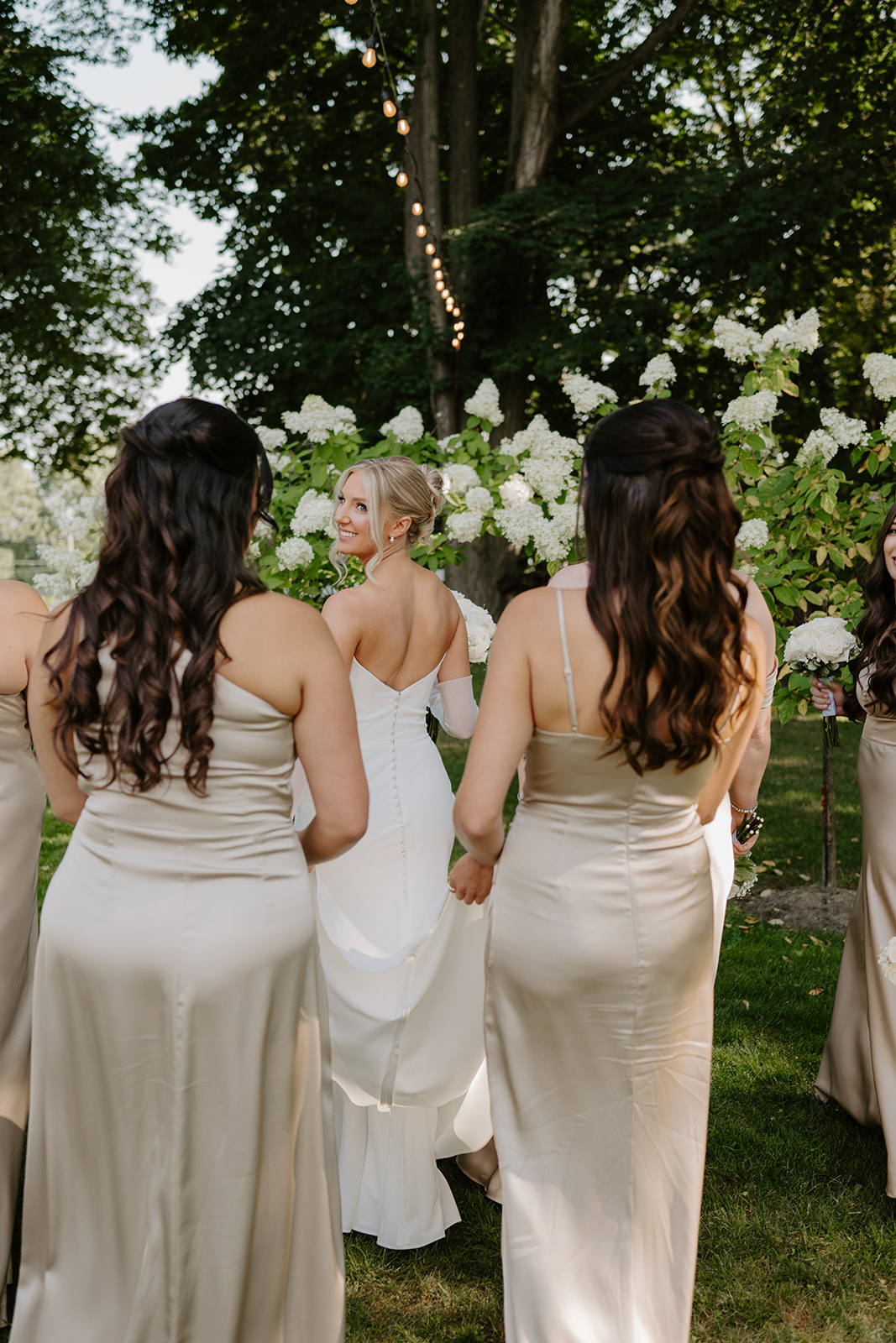 Bride looks back with a joyful smile while walking through a garden with her bridesmaids.